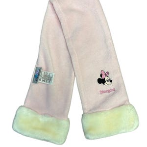 May include: A pale pink scarf with cream faux fur trim. The scarf has an embroidered Minnie Mouse design with a pink bow and the word "Disneyland" in pink script.