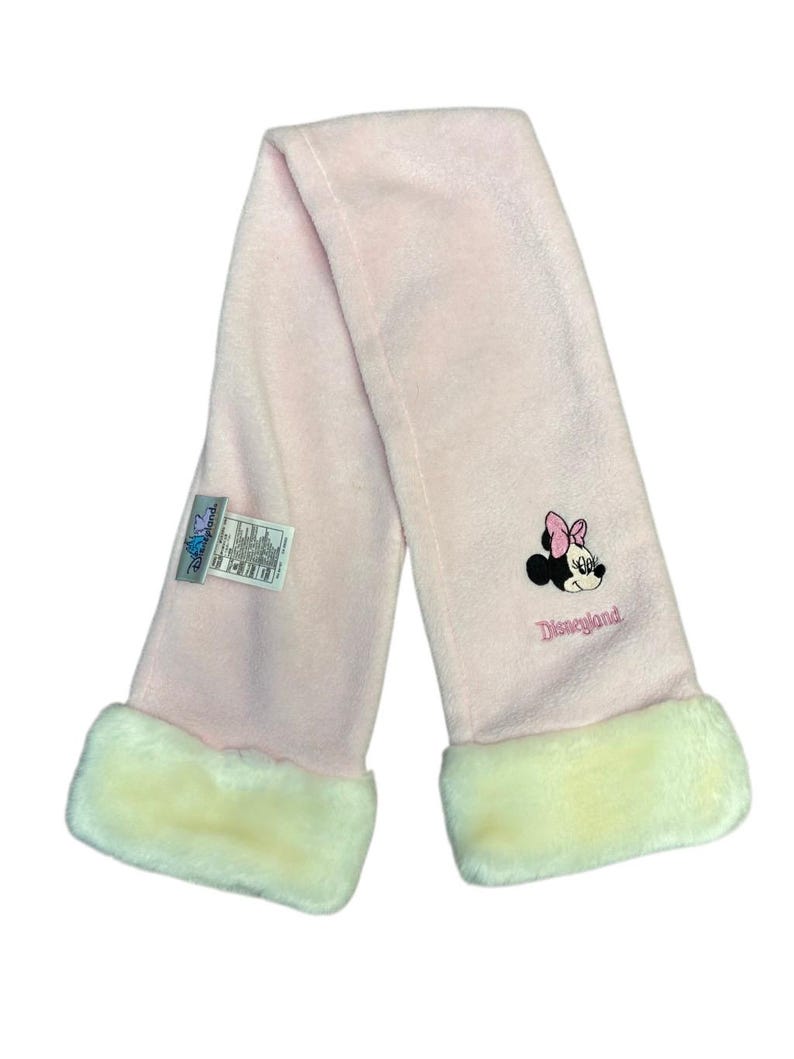 May include: A pale pink scarf with cream faux fur trim. The scarf has an embroidered Minnie Mouse design with a pink bow and the word "Disneyland" in pink script.