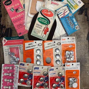 May include: A collection of vintage sewing supplies, including iron-on pockets, elbow patches, and various button kits. The items are packaged in colorful cardboard with text and illustrations, showcasing a retro aesthetic.