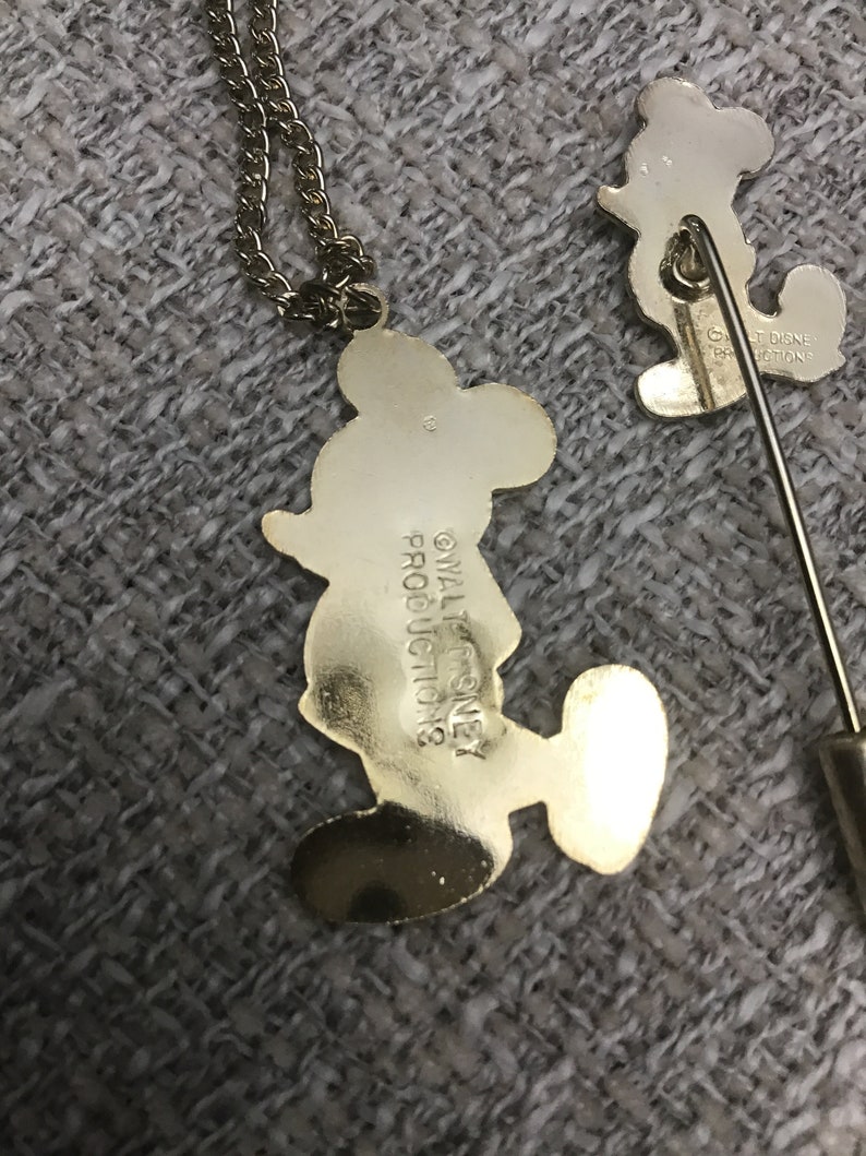 Puede incluir: Un conjunto de collar y broche de Mickey Mouse en tono dorado. El collar presenta un colgante de Mickey Mouse con el texto "&copy; WALT DISNEY PRODUCTIONS" en la parte posterior. El broche presenta un dise&ntilde;o de Mickey Mouse con el texto "&copy; WALT DISNEY PRODUCTIONS" en la parte posterior.