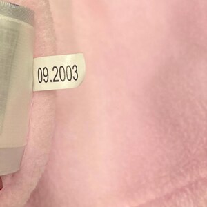 May include: A close-up of a soft, pink fabric with a tag attached. The tag displays the date "09.2003" in black text. The fabric appears to be plush and textured, with a light pink hue.