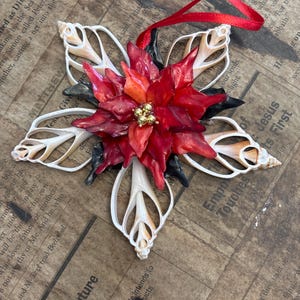 May include: A star-shaped Christmas ornament. It features a central poinsettia made of red and orange petals with gold accents. The star's points are formed from white seashells. It has a red ribbon for hanging.