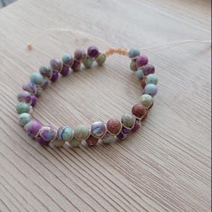 Peut inclure: Un bracelet fait à la main avec des perles rondes multicolores dans des tons de violet, vert et marron. Les perles sont enfilées avec un cordon beige, créant un accessoire unique. Le bracelet est présenté sur une surface en bois clair.