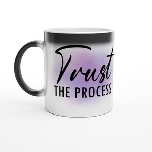Op de afbeelding: Een witte keramische koffiemok met een zwarte handgreep. De mok heeft een galaxie-achtig ontwerp met paarse en roze tinten. De tekst "Trust the Process" is in zwart op de mok gedrukt.