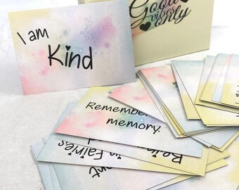Puede incluir: Un juego de cartas de afirmación con un fondo de acuarela de arcoíris pastel y detalles de purpurina. Las cartas presentan mensajes positivos como "Soy amable" y "Recuerda tu memoria". Las cartas vienen en una caja con las palabras "Good vibes only" en la tapa.