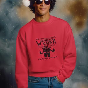 Könnte beinhalten: Rotes Sweatshirt mit dem Text "Gangsta Wrappa" und einer Geschenk-Grafik. Die Person im Bild trägt einen Weihnachtsmannhut und eine Sonnenbrille. Das Sweatshirt wird mit einer blauen Jeans kombiniert.