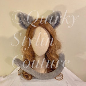 Może przedstawiać: Szary zestaw opaski na uszy kota i ogona z imitacji futra. Uszy są wyprostowane i puszyste, a ogon zakrzywiony. Zestaw jest prezentowany na głowie manekina z jasnobrązowymi włosami. Widoczny jest tekst "Quirky Stylin Couture".
