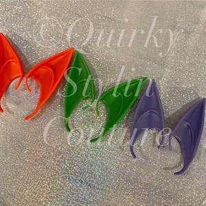 May include: Five pairs of elf ears in different colours: orange, green, purple. The ears are made of a flexible material and are designed to be worn on the head.