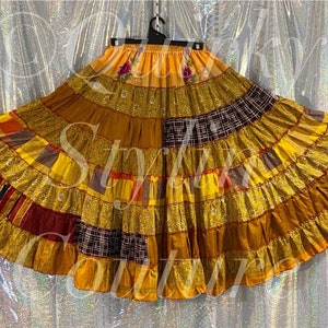 Könnte beinhalten: Ein mehrlagiger Patchwork-Rock in Gold-, Gelb- und Brauntönen. Der Rock weist eine Vielzahl von Stoffen auf, darunter schimmerndes Goldmaterial und gemusterte Quadrate. Der Bund ist einfarbig gelb, und der Rock hängt an einem schwarzen Kleiderbügel.
