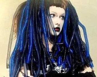 Könnte beinhalten: Eine Person mit dunklem Haar, das in zwei hohen Zöpfen gestylt ist, verziert mit blauen und schwarzen Schläuchen und schwarzem Netz. Die Person trägt dunkles Make-up, einschließlich dunklem Lippenstift, und ein schwarzes Oberteil.