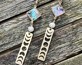 May include: A pair of gold-tone drop earrings featuring a square abalone shell inlay, a single white pearl, and a rectangular gold-tone pendant with a cutout moon phase design hanging from a standard earring hook.
