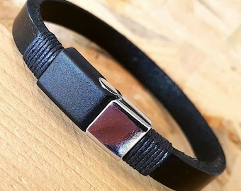 May include: A black leather bracelet featuring a smooth, rectangular silver-toned metal clasp. The leather band is secured to the clasp with dark, tightly wound thread detailing on both sides, creating a clean and structured appearance.