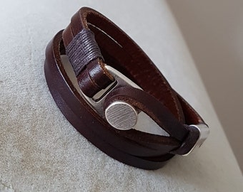 May include: A dark brown leather wrap bracelet featuring a brushed silver-tone metal circular clasp and a decorative dark brown cord wrapping detail. The bracelet consists of two leather bands that overlap and secure with the metal hardware.