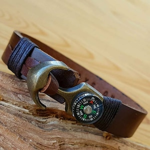 May include: A brown leather bracelet with a compass and an anchor-shaped clasp. The compass has a black face with white markings and a red needle.
