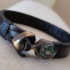 May include: Black leather bracelet with a compass and anchor clasp. The bracelet features a small compass with green, white, and red markings. The anchor clasp is a bronze color.