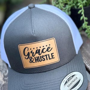 May include: A gray and white trucker hat with a leather patch that reads "Grace & Hustle" in black script. The hat has a curved brim and a snapback closure. The hat is a Yupoong Snapback.