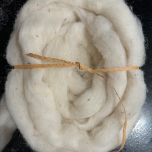 May include: A tightly rolled bundle of off-white wool roving, secured with a thin, light brown raffia tie. The wool has a soft, textured appearance, with subtle variations in color and small flecks throughout. The bundle is presented against a dark background.