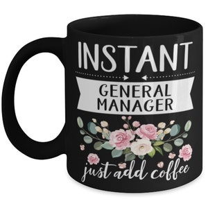 May include: Black coffee mug with white text that reads "Instant General Manager just add coffee". The text is surrounded by a white banner with arrows on either side. The mug is decorated with pink and green floral designs.