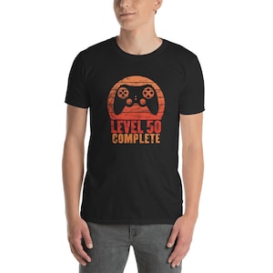 May include: Black t-shirt with a retro-style design featuring a video game controller inside a sunset-colored circle. The text "LEVEL 50 COMPLETE" is printed below the controller in orange.
