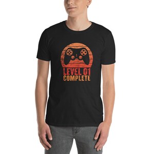 May include: Black t-shirt with a retro-style sunset graphic featuring a video game controller and the text "LEVEL 01 COMPLETE".