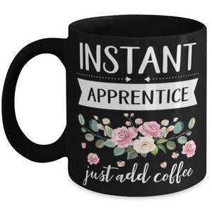 May include: Black coffee mug with white text that reads "Instant Apprentice just add coffee". The mug has a floral design with pink and white flowers and green leaves.