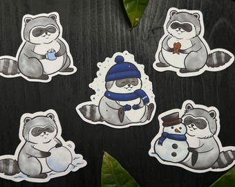 Op de afbeelding: Set van vijf wasbeerstickers in verschillende winterse scènes. Eén wasbeer houdt een mok vast, een ander een koekje, en een derde bouwt een sneeuwpop. De andere twee wasberen dragen een hoed en sjaal.