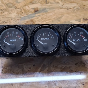 Può includere: Tre indicatori neri con lancette bianche e segni rossi. Gli indicatori leggono "VOLT", "OIL/KG" e "OIL/T C".