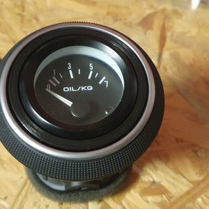 Könnte beinhalten: Ein schwarzes Öldruckmessgerät mit weißem Zifferblatt und Zeiger. Das Messgerät zeigt OIL/KG an und hat Markierungen von 0 bis 5. Das Messgerät hat eine strukturierte schwarze Lünette und einen silbernen Zierring.