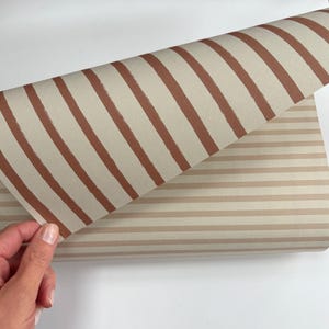 May include: A roll of wallpaper featuring two distinct patterns. The top layer displays diagonal, thick, hand-painted style terracotta stripes on a light beige background, while the bottom layer shows horizontal, thin, light brown stripes.