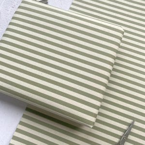 May include: Sage green and white striped wrapping paper, neatly folded into a stack. A sheet of the same pattern lies beside it. A pair of silver scissors is visible in the bottom right corner.