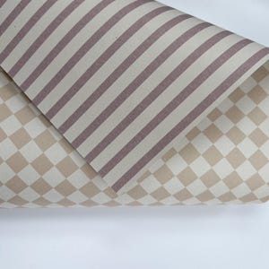 May include: A sheet of material featuring two distinct patterns. The top layer displays a series of diagonal mauve and cream stripes, while the bottom layer shows a repeating cream and beige checkered pattern across the surface of the material.