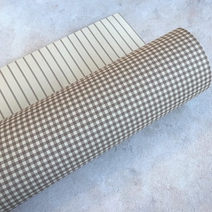 May include: A roll of fabric featuring a brown and white gingham check pattern, partially unrolled to reveal an underlying layer of off-white fabric with thin, vertical brown stripes running parallel along the length of the material.