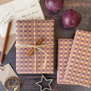 May include: A still life composition with wrapped gifts in a brown and tan checkered pattern, adorned with raffia and cinnamon sticks. The scene also includes plums, stationery, and a star-shaped cookie cutter. The color scheme is warm and earthy.