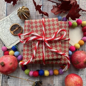 May include: A wrapped present with red and white twine, featuring a red and tan chequered pattern. The gift is surrounded by colourful felt ball garland, pomegranates, dried leaves, and a map on a wooden surface.