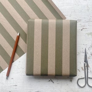 May include: A wrapped gift box with olive green and beige vertical stripes, a matching sheet of wrapping paper, a brown pencil, and small silver scissors. The items are arranged on a distressed white wooden surface.