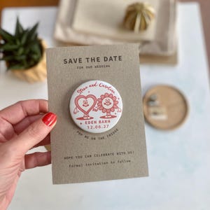 May include: Save the date card with a round button featuring a cartoon heart and flower holding hands. The button says "Steve and Caroline" and "Eden Barn 12.06.27". The card says "Pop me on the fridge" and "Hope you can celebrate with us! Formal invitation to follow."