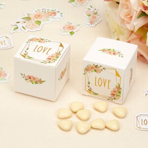 Puede incluir: Dos cajas de regalo blancas con diseños florales y detalles dorados. Las cajas tienen la palabra "LOVE" impresa. Las cajas están rodeadas de caramelos blancos en forma de corazón.