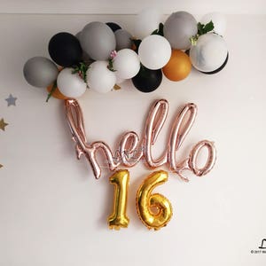 Puede incluir: Globos de lámina de oro rosa que deletrean "Hello 16" con globos de números dorados. Los globos están rodeados por una guirnalda de globos negros, blancos y dorados.