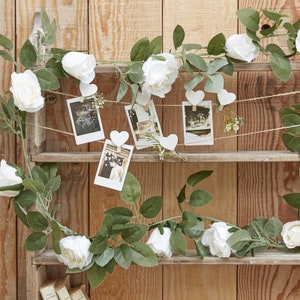 May include: White faux flower garland with white heart-shaped clothespins holding up instant photos on a wooden shelf.