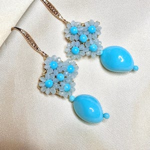 Peut inclure: Paire de boucles d'oreilles pendantes avec un motif floral, des perles bleu clair et des pendentifs turquoise en forme de goutte. Les boucles d'oreilles ont des crochets d'oreilles dorés et sont présentées sur une surface crème.