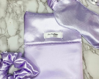 Könnte beinhalten: Ein lavendelfarbenes Schlafset aus Satin, bestehend aus einem rechteckigen Beutel, einem Haargummi und einer Schlafmaske. Der Beutel und die Maske haben ein weißes Stoffetikett mit dem schwarzen Text "Kila May Designs".