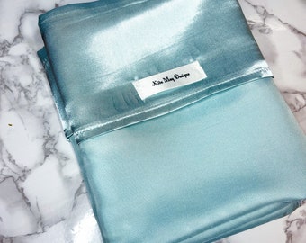 Könnte beinhalten: Ein gefalteter hellblauer Satin-Kissenbezug mit glatter, reflektierender Oberfläche. Ein weißes, rechteckiges Stoffetikett ist am oberen Rand aufgenäht und trägt den schwarzen Aufdruck Kita May Designs in einer klaren Schrift.