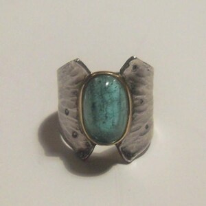 May include: A silver ring with a textured design and a large oval-shaped turquoise gemstone set in a gold bezel.