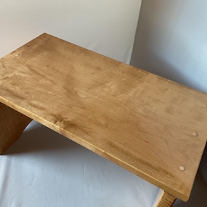 May include: A light brown wooden step stool with a rectangular top and angled legs. The stool is made of light-colored wood and has a smooth, polished finish. The stool is approximately 24 inches long, 12 inches wide, and 10 inches tall.