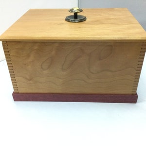 May include: A wooden box with a cherry wood finish and a brass knob on the lid. The box has a red wood trim around the bottom.