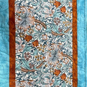 Op de afbeelding: Een turquoise en oranje tafelkleed met een bloemenpatroon. De stof heeft een herhalend ontwerp van gestileerde lelies en bladeren. Het tafelkleed is 39,4 cm x 97,8 cm en is gemaakt van katoen met polyester vulling.