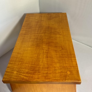 Op de afbeelding: Een houten kast met een goudbruin, getextureerd bovenblad. De houtnerf is zichtbaar en creëert een golvend patroon. De kast heeft een eenvoudig, rechthoekig ontwerp, met een lichtgekleurde basis. De bovenkant is afgewerkt met een glanzende vernis.