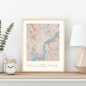 May include: A framed print of a map of Philadelphia, Pennsylvania. The map is in shades of brown, blue, and gray. The city name is printed in a colorful font at the bottom of the map.