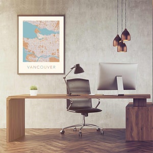Op de afbeelding: Een ingelijste kaartprint van Vancouver in beige, oranje en blauwe tinten hangt boven een houten bureau. Het bureau heeft een zwarte bureaustoel, een computermonitor en een kleine plant in een pot. Een zwarte bureaulamp en drie hanglampen maken de kantoorinrichting compleet.