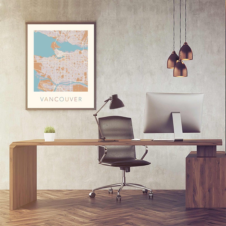 Op de afbeelding: Een ingelijste kaartprint van Vancouver in beige, oranje en blauwe tinten hangt boven een houten bureau. Het bureau heeft een zwarte bureaustoel, een computermonitor en een kleine plant in een pot. Een zwarte bureaulamp en drie hanglampen maken de kantoorinrichting compleet.